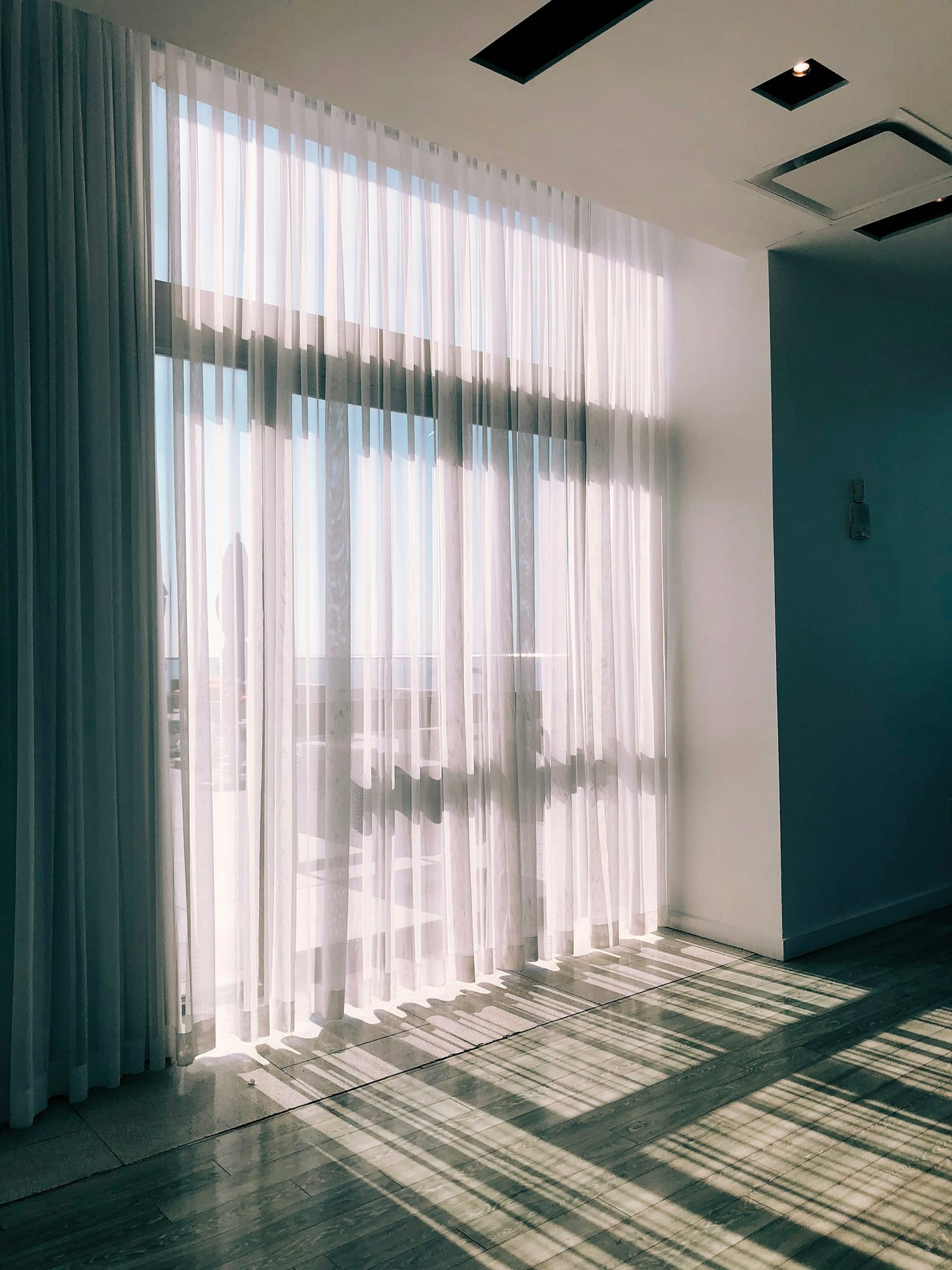 Sunlight through floor-to-ceiling sheer curtains, casting dramatic shadow patterns